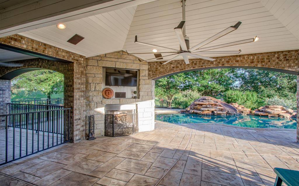 Spacious outdoor patio featuring a stone gas fireplace and a large Big Ass fan. It opens up to beautiful pool area with rock formations, surrounded by lush greenery, offering a perfect space for relaxation and entertainment.