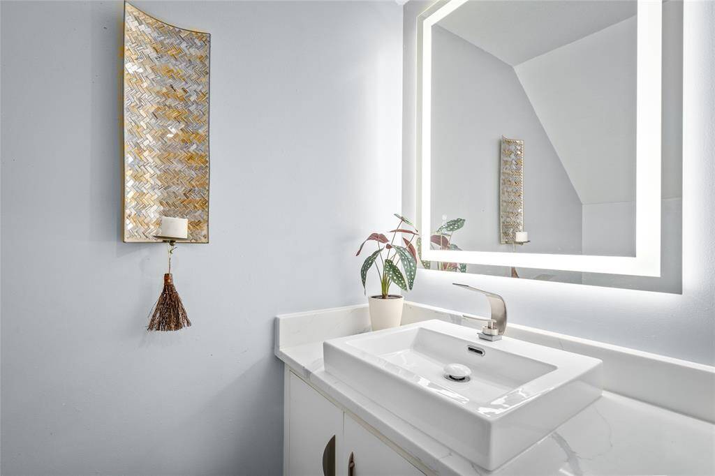 This elegant downstairs half bath features a modern vessel sink atop a sleek quartz countertop. A backlit mirror adds a contemporary touch, while the neutral tones and decorative wall accents create a warm and inviting space. Perfect for guests, this thoughtfully designed powder room combines function and style.