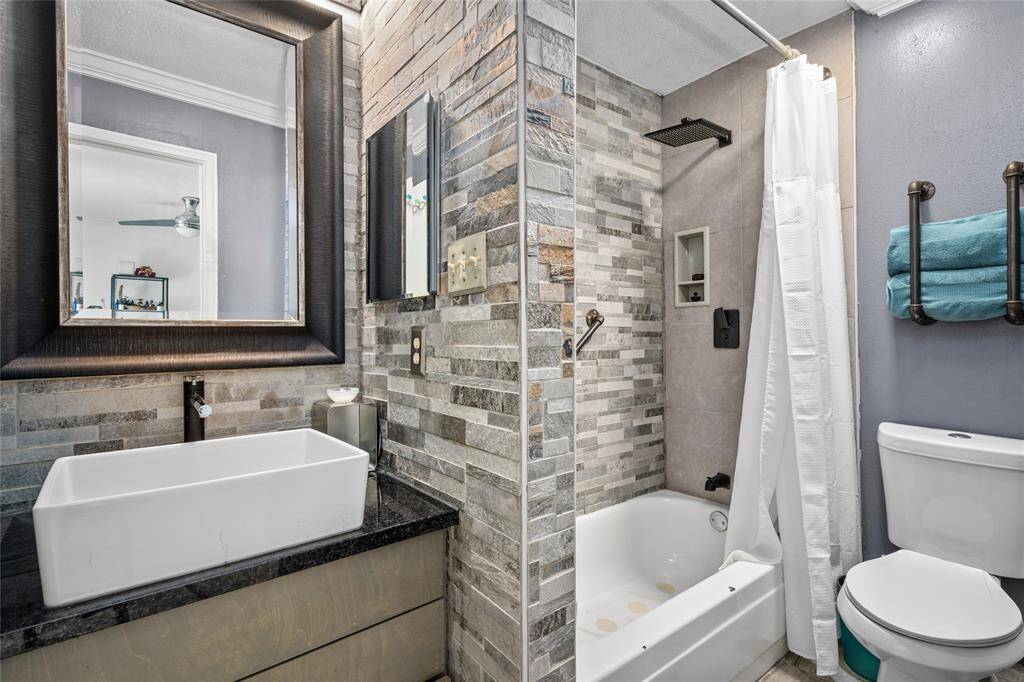 This beautifully updated primary bathroom features a modern, spa-like aesthetic with a striking stone accent wall and a sleek black granite vanity. The vessel sink and framed mirror add a touch of sophistication, while the walk-in shower boasts a rainfall showerhead for a luxurious bathing experience. A deep soaking tub provides an additional option for relaxation. Thoughtful storage solutions, including built-in shelving and industrial-style towel racks, enhance both style and functionality.