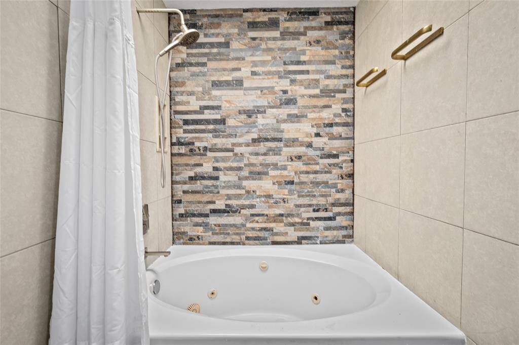 Unwind in the luxurious primary bath featuring a relaxing jetted tub, perfect for soaking after a long day. The stunning accent wall with modern stacked stone tile adds a touch of elegance, while the handheld showerhead provides both convenience and style. Sleek, neutral-toned tiles and gold-tone towel racks complete this spa-like retreat.