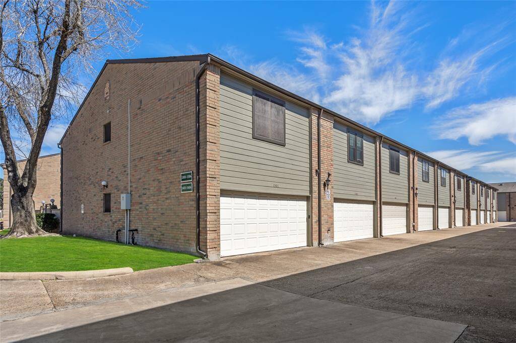 Rear view of the well-maintained townhome community, showcasing a row of two-car garages with clean white doors. The brick and siding exterior provides a classic and modern blend, while upper-level windows invite natural light inside. The paved driveway offers ample space for parking and easy access. A neatly landscaped green space with a mature tree adds a touch of nature to the area.