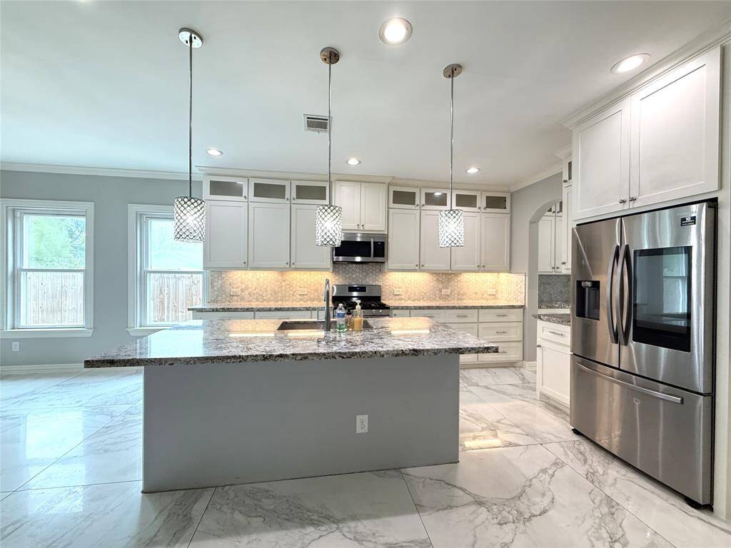 This modern kitchen features a spacious island with granite countertops, stainless steel appliances, and sleek white cabinetry. It has elegant pendant lighting and large windows offering plenty of natural light. The room is finished with polished marble-style flooring for a clean, sophisticated look.