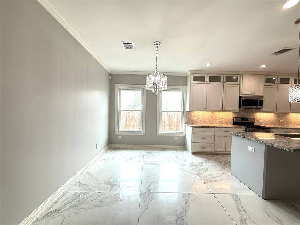 This photo features a modern kitchen and breakfast area with large marble-like tile flooring. The kitchen has white cabinetry, granite countertops, and stainless steel appliances, including a built-in microwave. Two windows provide natural light, and elegant pendant lighting adds a stylish touch.
