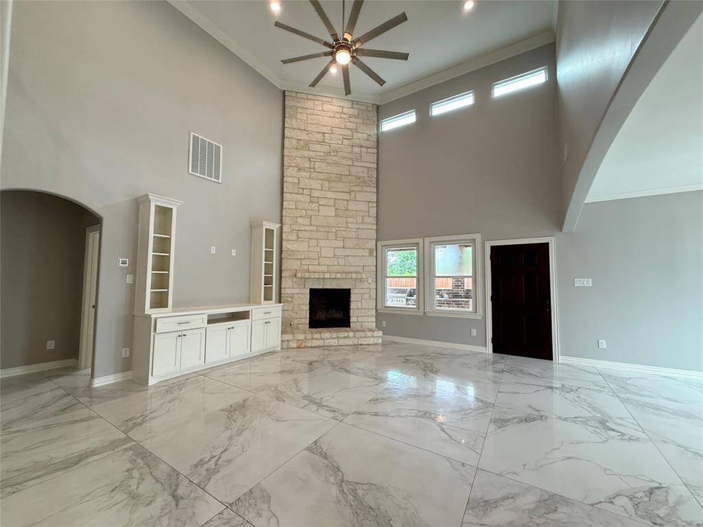 This spacious living room features high ceilings, a modern ceiling fan, and a stunning stone fireplace. It includes built-in shelving, large windows for natural light, and elegant marble-like tile flooring, creating a bright and inviting atmosphere.