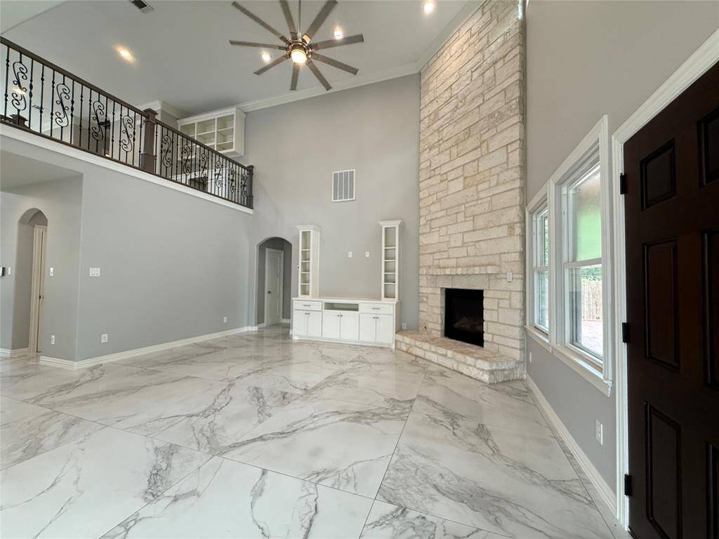 Spacious living room with high ceilings, large stone fireplace, and elegant marble-like flooring. Features built-in shelving and abundant natural light through upper windows, creating a bright and inviting space.
