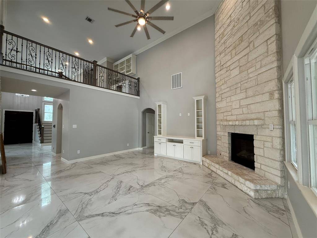 This spacious living area features high ceilings, a large stone fireplace, elegant marble-like flooring, and a wrought iron staircase. The room is bright with neutral tones and has built-in cabinets for storage.
