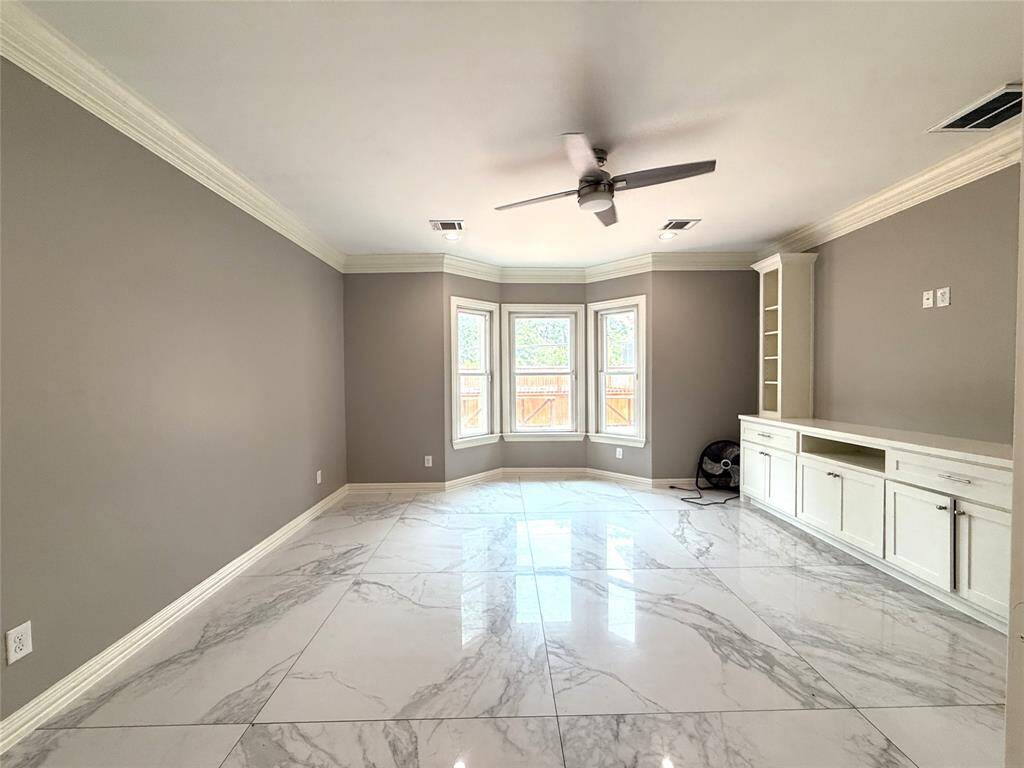 Bright, spacious master bedroom with large windows, marble-style tiled floor, neutral gray walls, and white built-in cabinets. Features a ceiling fan and crown molding for a modern touch.