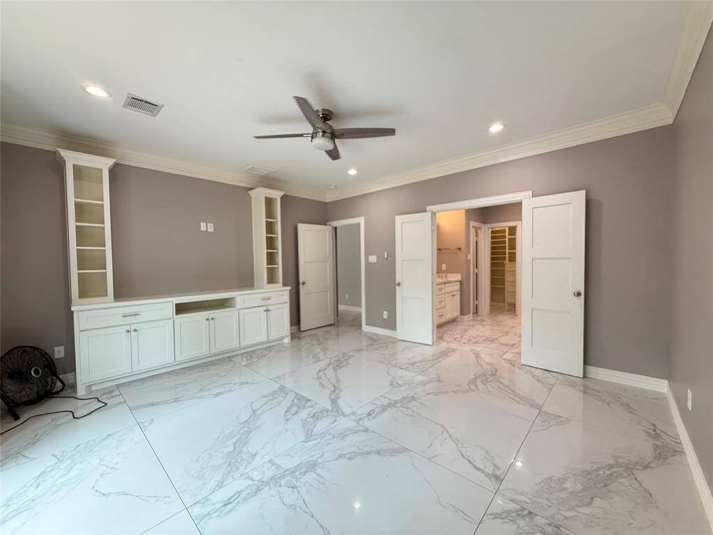Spacious master bedroom with elegant marble-like tile flooring and neutral gray walls. Features built-in shelving and cabinetry, a ceiling fan, recessed lighting, and an ensuite bathroom with a walk-in closet.