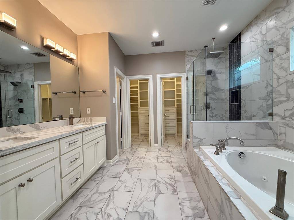 This master bathroom features elegant marble flooring and soft gray walls, complemented by built-in white cabinetry and shelving. It includes recessed lighting, a ceiling fan, and opens to a spacious bathroom and walk-in closet through double doors, offering a sleek and modern design.