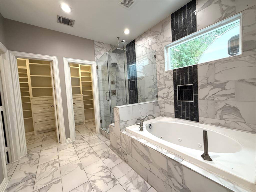 This spacious bathroom features elegant marble tile, a large soaking tub, a glass-enclosed shower, and dual walk-in closets with built-in storage, offering both luxury and functionality.