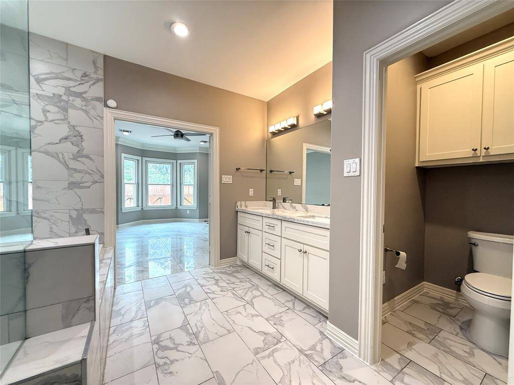 This photo showcases a modern bathroom with elegant marble tile flooring and walls, a double vanity with ample storage, and a separate toilet area. The space is brightened by recessed lighting and adjoins a room with large bay windows and a ceiling fan.