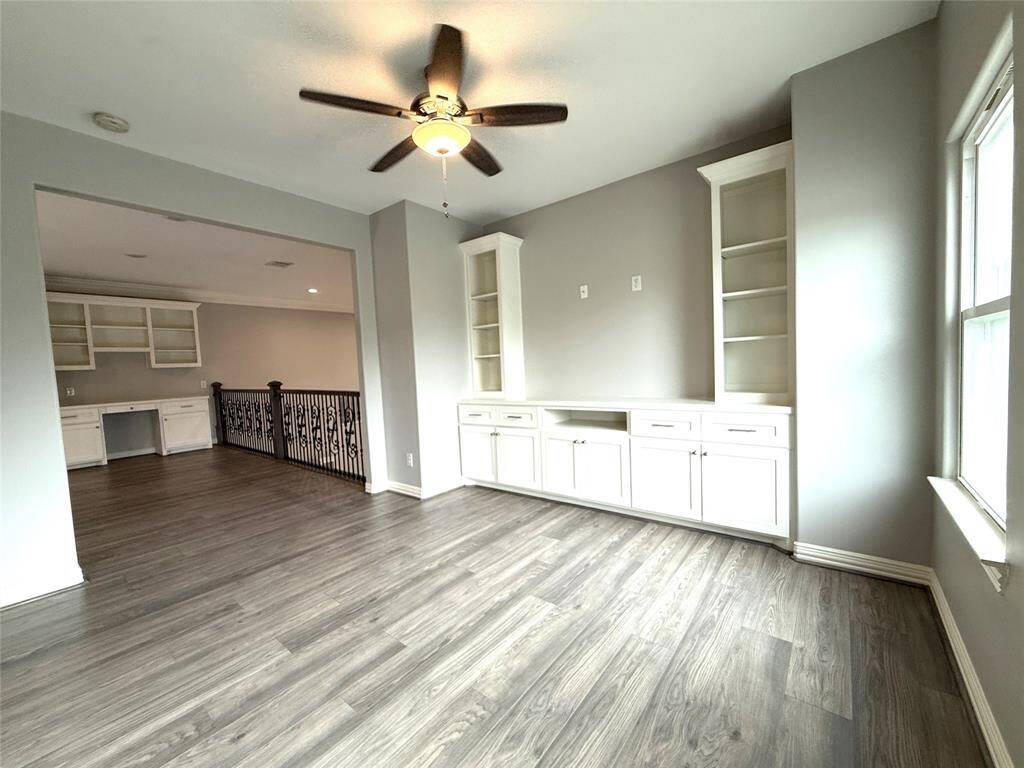 This room features modern gray flooring, built-in white cabinetry, and ample natural light from large windows. A ceiling fan adds comfort, while the open layout connects to an adjacent space with additional cabinetry and decorative railing. Ideal for a bright, functional living area.