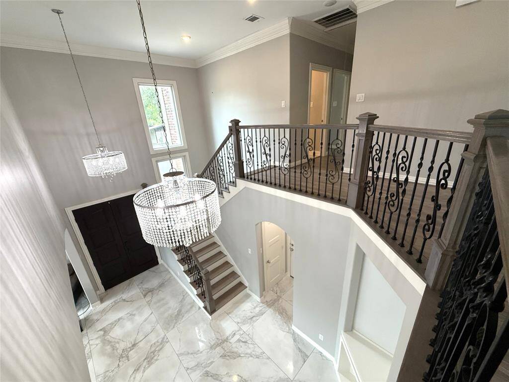 This photo showcases a spacious, elegant foyer with high ceilings, a grand staircase featuring ornate wrought iron railings, and beautiful chandelier lighting. The area has a modern, open feel with large, glossy marble-like tiles and ample natural light from tall windows.
