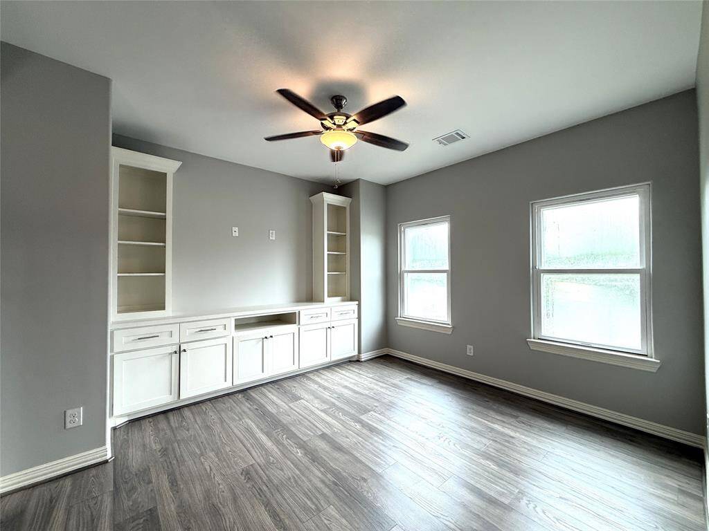 This room features sleek hardwood floors, built-in white cabinetry with shelving, two large windows for natural light, and a ceiling fan for comfort.
