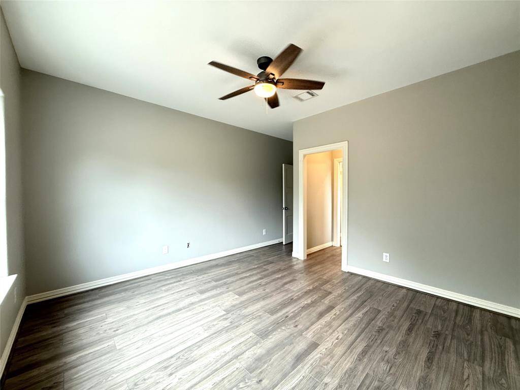 Bright, spacious room with modern wood flooring, neutral walls, and a ceiling fan. Natural light floods in through a large window, creating a welcoming atmosphere.