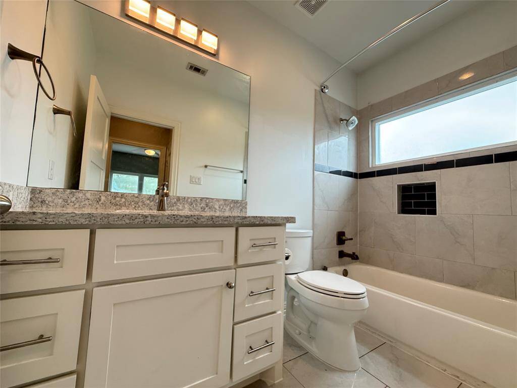 This bathroom features a modern design with a granite countertop, under-mount sink, and ample cabinetry. It includes a bathtub with a sleek tile surround, a large mirror, and a window for natural light.