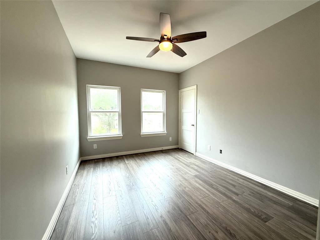 Bright, spacious room with light gray walls and wood-style flooring. Features a ceiling fan and two large windows providing natural light.