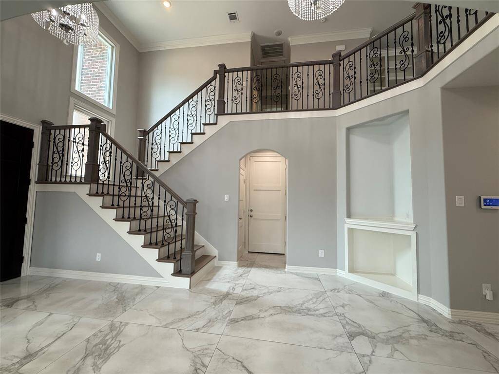 This elegant entryway features a grand staircase with ornate iron railings, high ceilings, and large windows that fill the space with natural light. The marble-like tile flooring and modern chandelier add a touch of luxury.