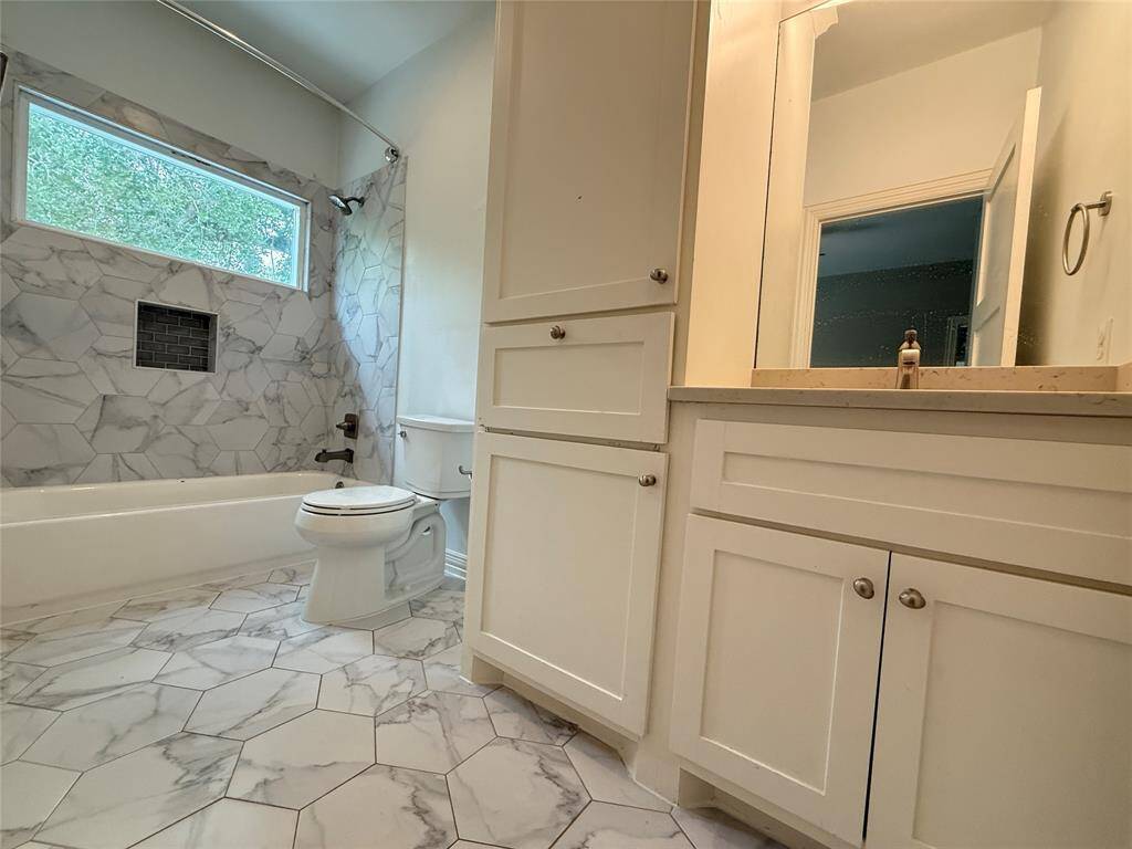 This bathroom features a modern design with marble-patterned tile flooring and walls, a large bathtub with a shower, a window for natural light, and ample storage with white cabinetry.