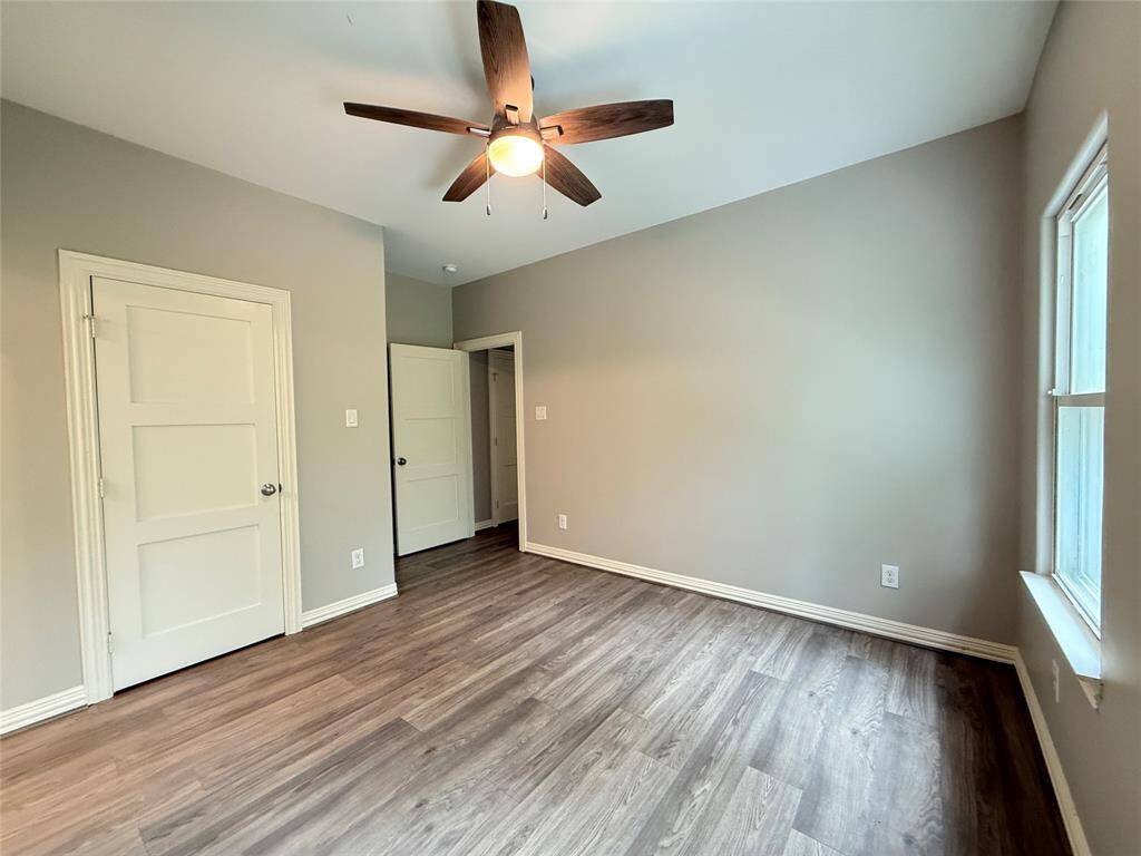 This room features a neutral color palette with light gray walls and wood-style flooring. It includes a ceiling fan with a light fixture, a window providing natural light, and two white doors, one of which is open to a hallway.
