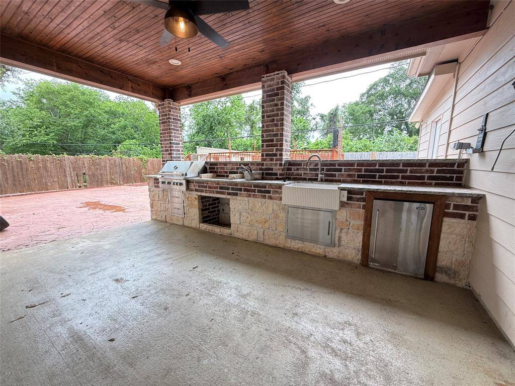 This photo showcases a spacious covered patio with a built-in outdoor kitchen featuring a grill, sink, and refrigerator. The area has a rustic brick and stone design, complemented by a wood-paneled ceiling with a ceiling fan, offering a great space for outdoor entertaining.