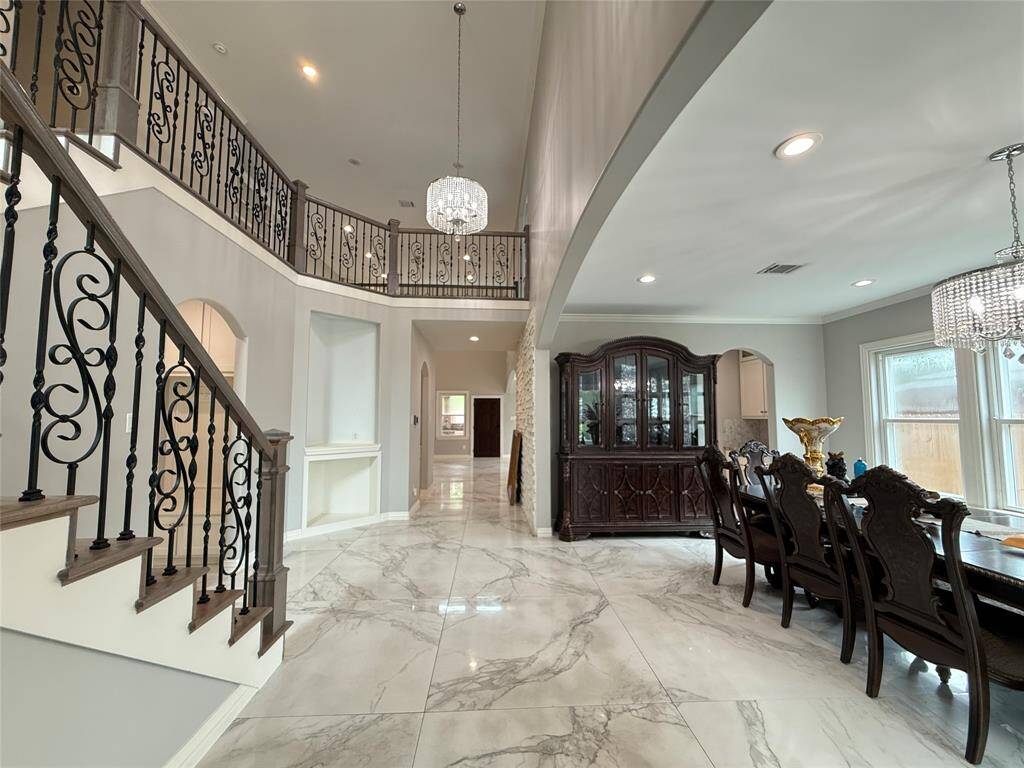 This spacious home features a grand entryway with elegant marble floors and a sweeping staircase. The open layout includes a formal dining area with large windows and ornate chandeliers, complemented by decorative iron railings and a sophisticated color palette.