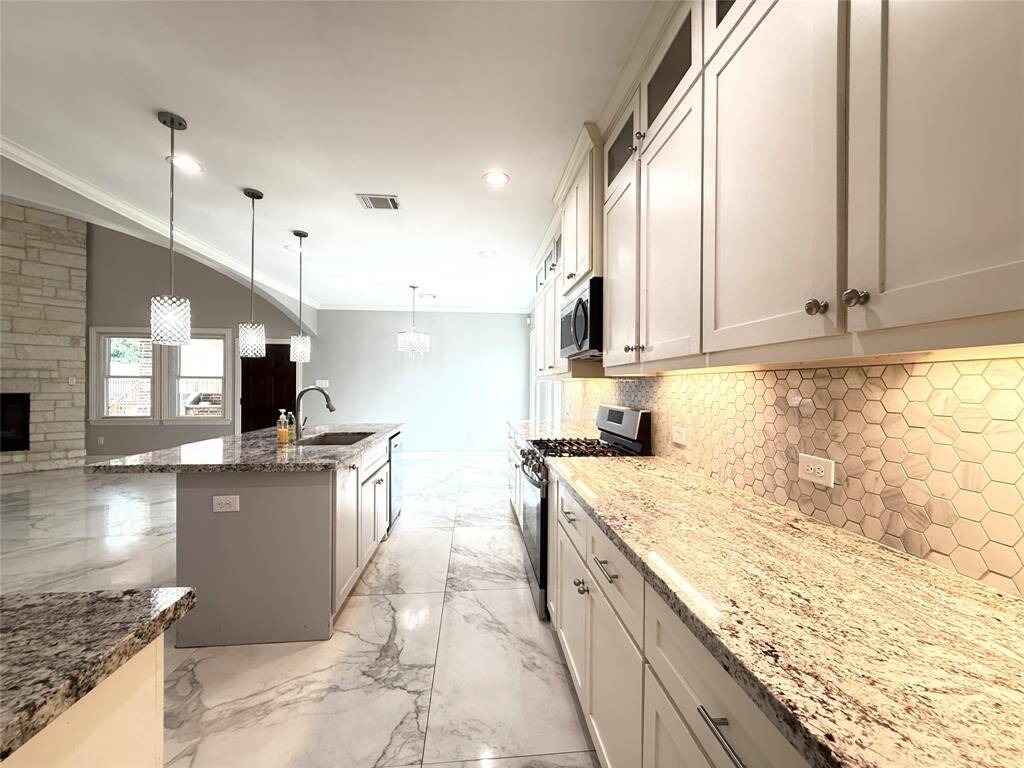 This photo showcases a spacious, modern kitchen with granite countertops, white cabinetry, and a large island. The room features pendant lighting, a stone backsplash, and an open layout with a view of a living area with a stone fireplace.