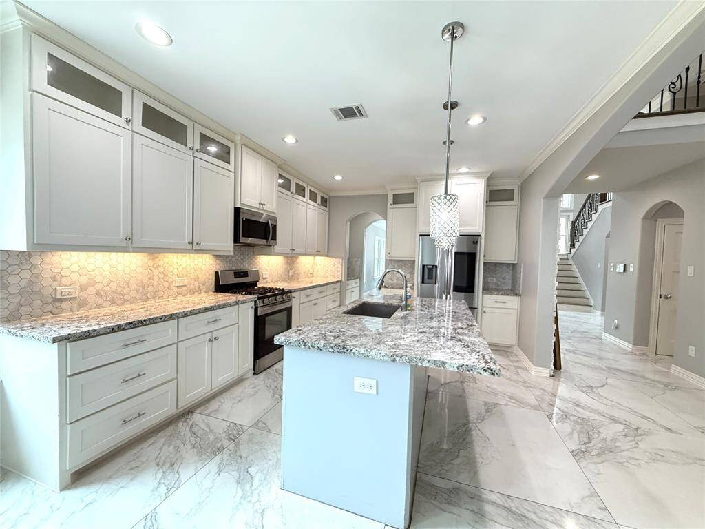 This kitchen features sleek white cabinetry, granite countertops, and modern stainless steel appliances. The spacious island offers extra prep space and seating, while the hexagonal tile backsplash adds a stylish touch. Bright and airy, with recessed lighting and marble-like tile flooring, it opens up to a staircase and additional living areas.