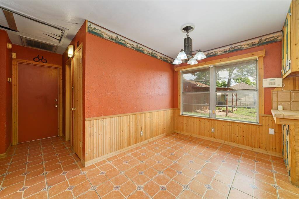 This room features warm terracotta tile flooring and wood paneling with a rustic red wall. It includes a large window for natural light and a hanging light fixture. The space offers access to the outdoors, ideal for a cozy dining area or sunroom.