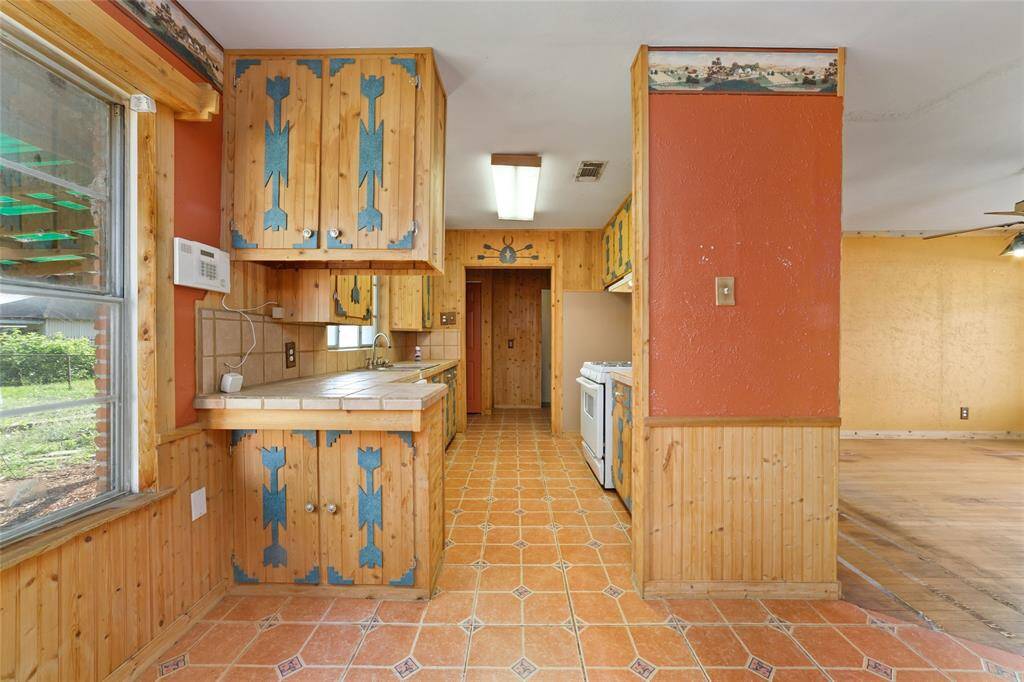 This cozy kitchen features rustic wood cabinetry with blue accents, tiled countertops, and a warm orange wall. It has a vintage feel with wood paneling and tiled floors, offering a unique charm and a view of the backyard.