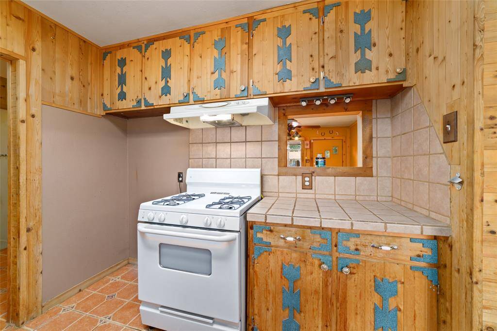 Cozy kitchen with a rustic charm featuring wooden cabinets with decorative blue accents, tiled countertops, and a gas stove. A small pass-through window adds character and connects to an adjacent room.