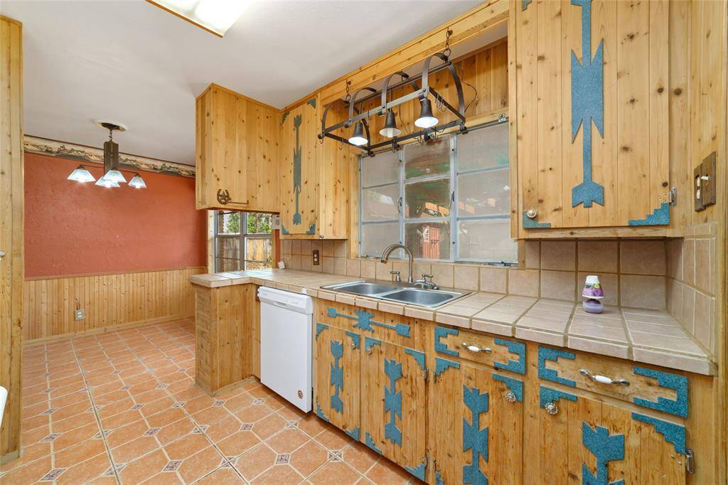 This photo showcases a rustic kitchen with distinctive wooden cabinets featuring blue decorative accents. It includes a dual sink set in a tiled countertop, a dishwasher, and a window above the sink. Adjacent is a dining area with warm-toned walls and matching tile flooring, complete with a vintage-style light fixture.