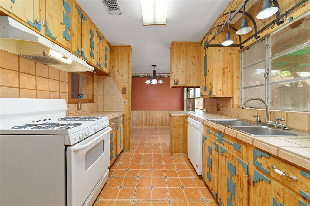 Cozy kitchen with a rustic feel, featuring wood cabinetry with blue accents, tile countertops, and a vintage gas stove. Ample natural light comes through large windows, enhancing the warm, earthy tones of the tiled floor.
