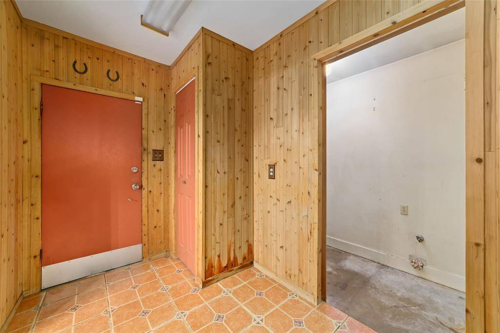 This photo shows a small entryway with wood paneling and terracotta tile flooring. It features a red door and a small closet, alongside an unfinished adjacent room. The space has a rustic feel.