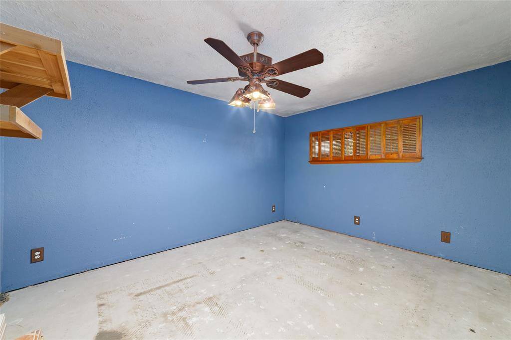 This room features blue walls, a ceiling fan with lights, and a wooden louvered window. The floor is bare, offering a blank canvas for flooring of your choice. There's also a small wooden shelf in the corner, adding a touch of functionality.