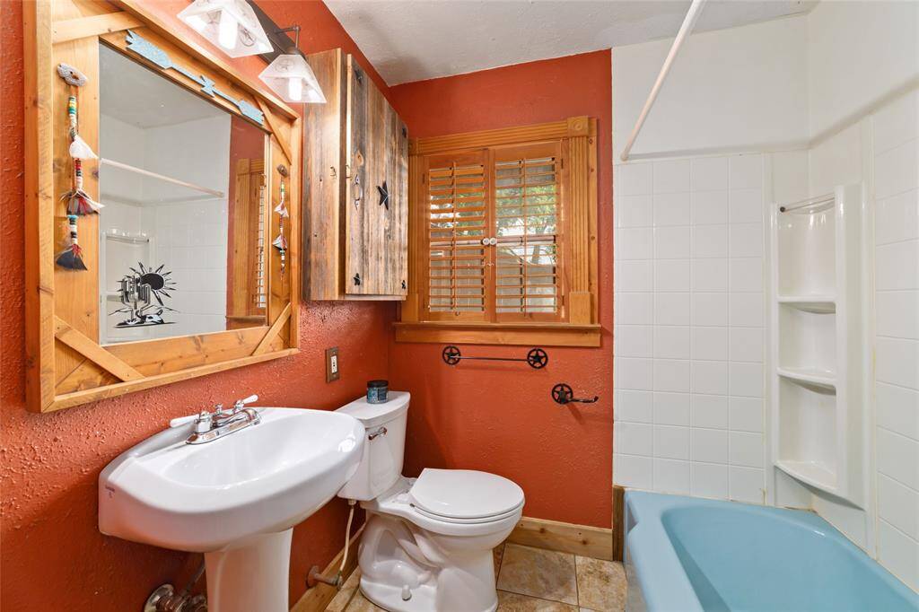 This cozy bathroom features a rustic style with warm terracotta walls and wood accents. It includes a pedestal sink, a toilet, and a bathtub with white tiled walls. A wooden cabinet and mirror add charm, while a window with shutters provides natural light.