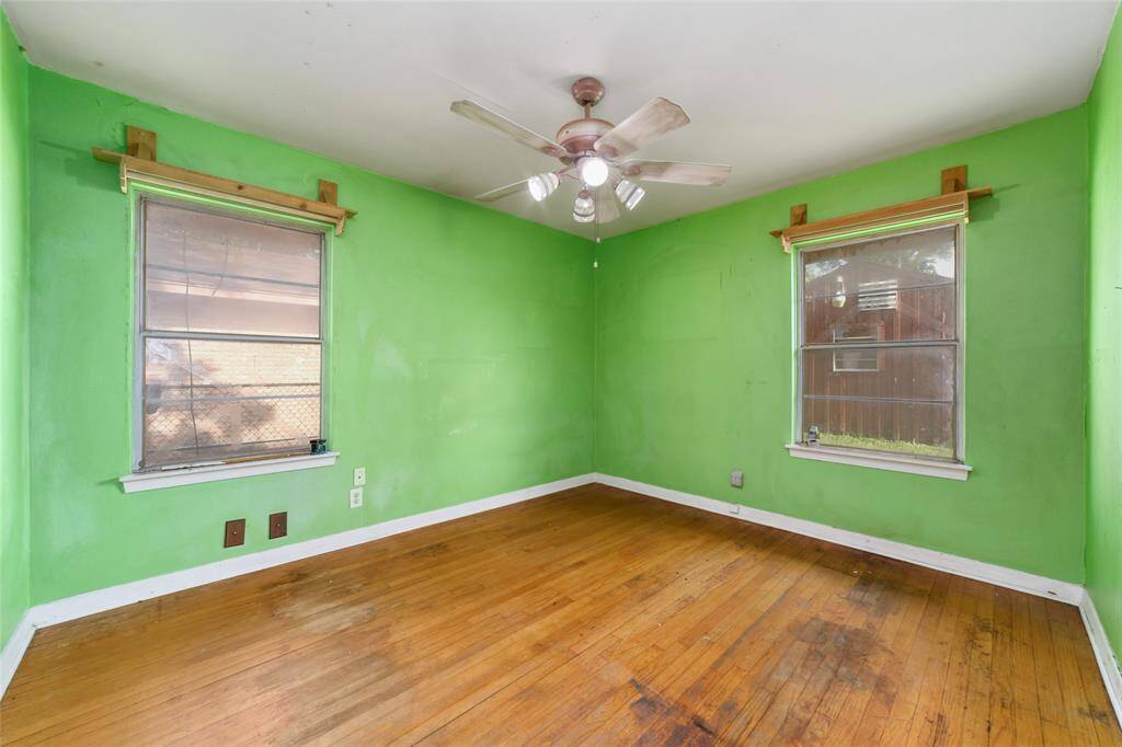Bright room with green walls, wood flooring, and two windows for natural light. Features a ceiling fan with lights, offering a cozy atmosphere. Some wear on the floor and walls suggests potential for personal touches.