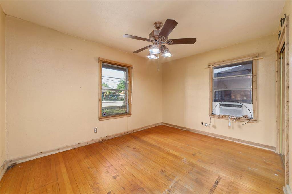 This room features hardwood flooring, two windows allowing natural light, a ceiling fan with lights, and an installed window air conditioner. It has simple, neutral-colored walls, offering a blank canvas for customization.