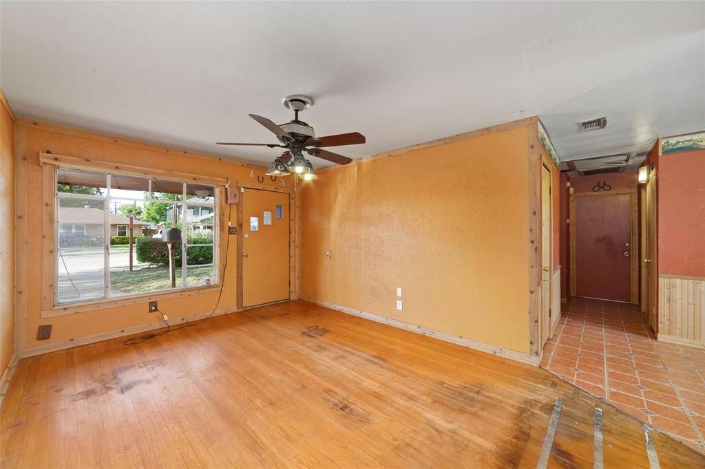 Cozy living area featuring hardwood floors and a large window providing natural light. Equipped with a ceiling fan, the space leads to a hallway with tiled flooring, offering potential for customization.