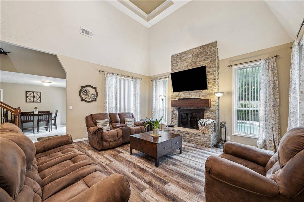 This inviting living room features a high ceiling, stone fireplace, and large windows for natural light. It has wood-style flooring creating a warm and welcoming atmosphere. The open layout leads to a dining area, enhancing the spacious feel.