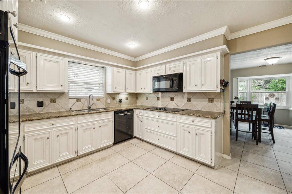 This kitchen features spacious white cabinetry, granite countertops, and a tile backsplash. It includes modern appliances, ample natural light from a large window, and opens to a dining area. The tile flooring completes the clean look.