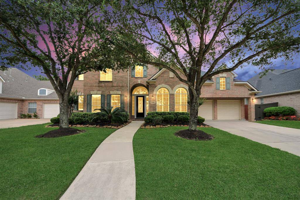 This inviting home features a brick exterior with large arched windows. It has a well-maintained front yard, a two-car garage, and a charming walkway leading to the entrance. Perfect for a family seeking comfort and style.
