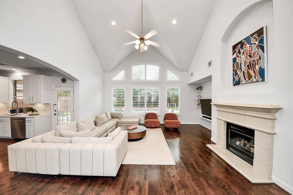 This spacious living room features high vaulted ceilings, large windows with abundant natural light, and a cozy fireplace. The open layout connects seamlessly to the modern kitchen, creating a welcoming and functional space for entertaining.