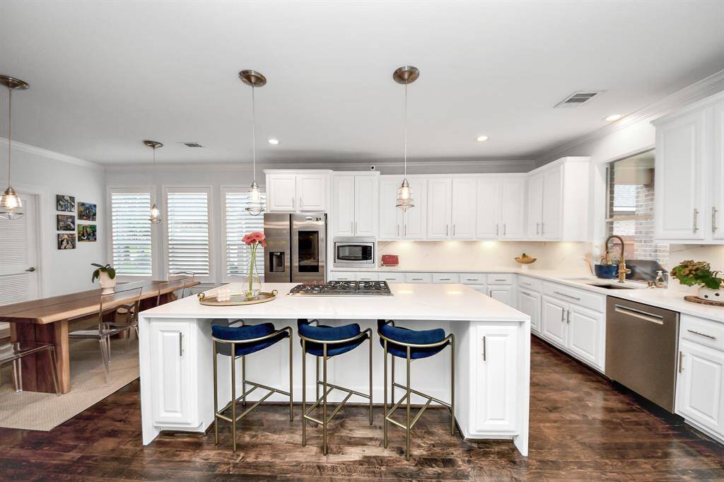 This kitchen features a spacious, modern design with white cabinetry, stainless steel appliances, and a large island with seating. Pendant lighting adds elegance, while hardwood floors extend to a dining area with natural light from large windows.