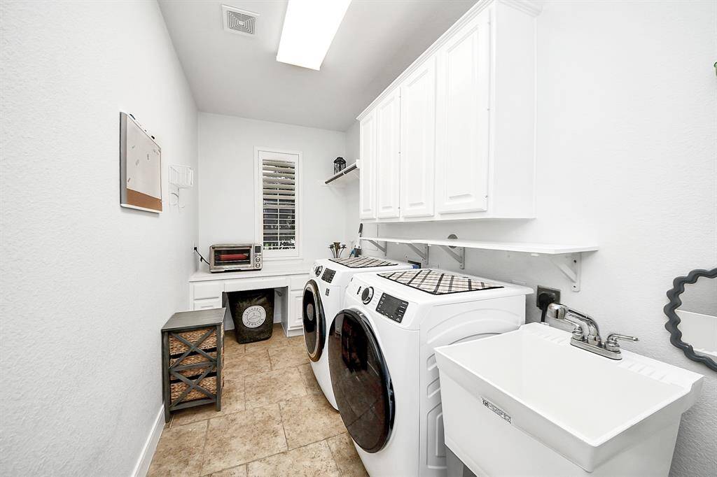 This is a bright, well-organized laundry room featuring a front-loading washer and dryer, ample white cabinetry for storage, a utility sink, and a small workspace with a window for natural light. The space is enhanced by tiled flooring and a clean, modern aesthetic.