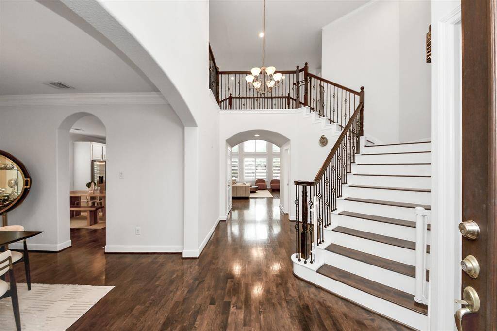 This elegant entryway features a grand staircase with wrought iron railings, hardwood floors, and high ceilings. Arched doorways lead to an open dining area and living space, creating a warm and inviting atmosphere.