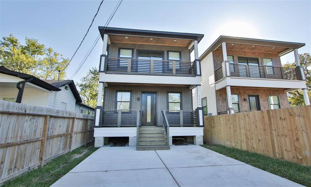 This modern two-story home features a sleek wood and black exterior, with two balconies and a welcoming entryway. The property includes a paved driveway and is surrounded by a wooden fence, offering privacy and a contemporary feel.