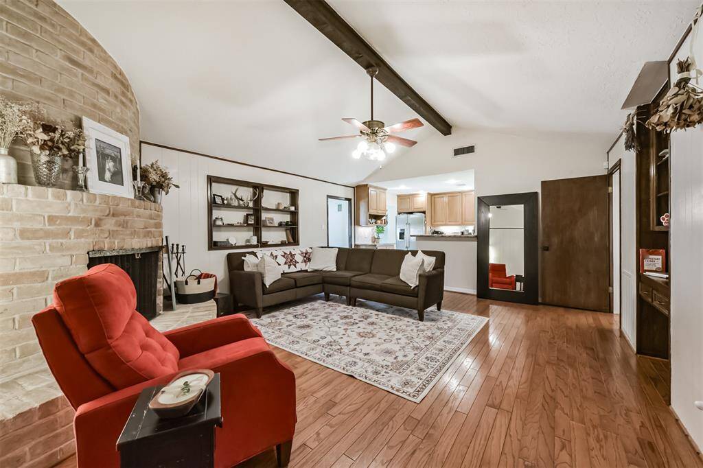 Spacious living area with wood floors, vaulted ceiling, brick fireplace, and built-in shelving. View looking into Kitchen