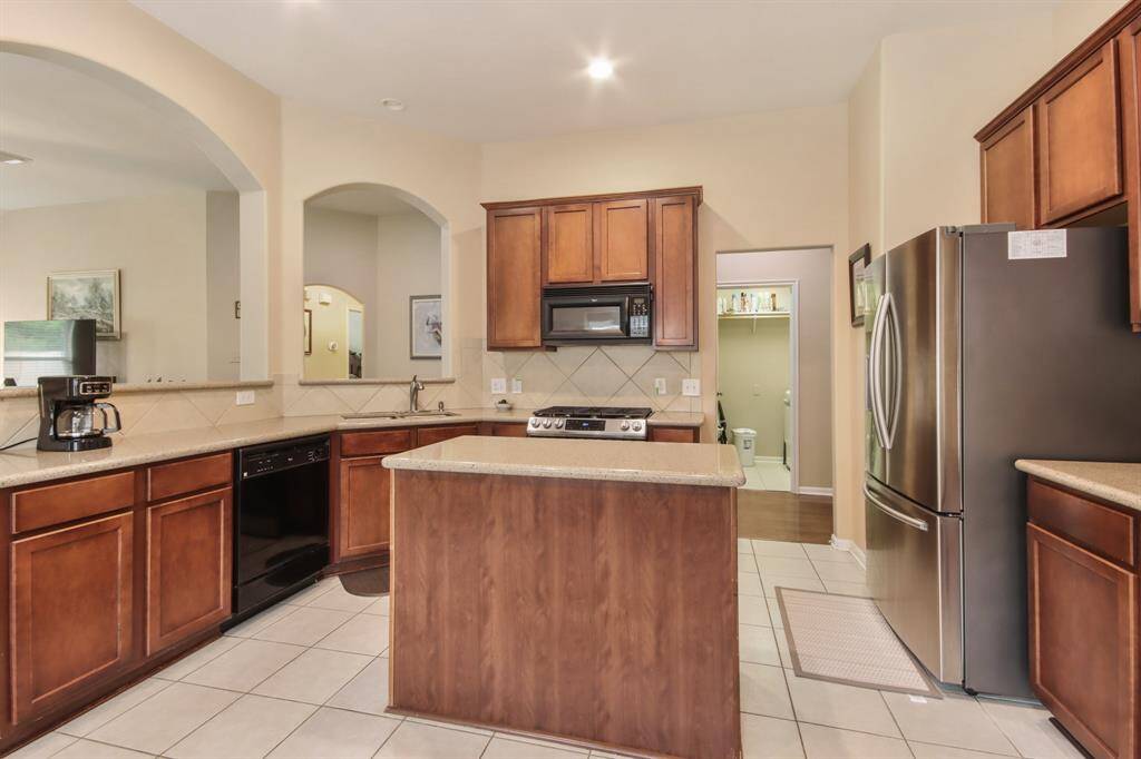 This kitchen features warm wood cabinetry, a central island, and modern stainless steel appliances. It has tiled floors and an open layout with arched doorways, offering a spacious and inviting atmosphere. Refrigerator included!
