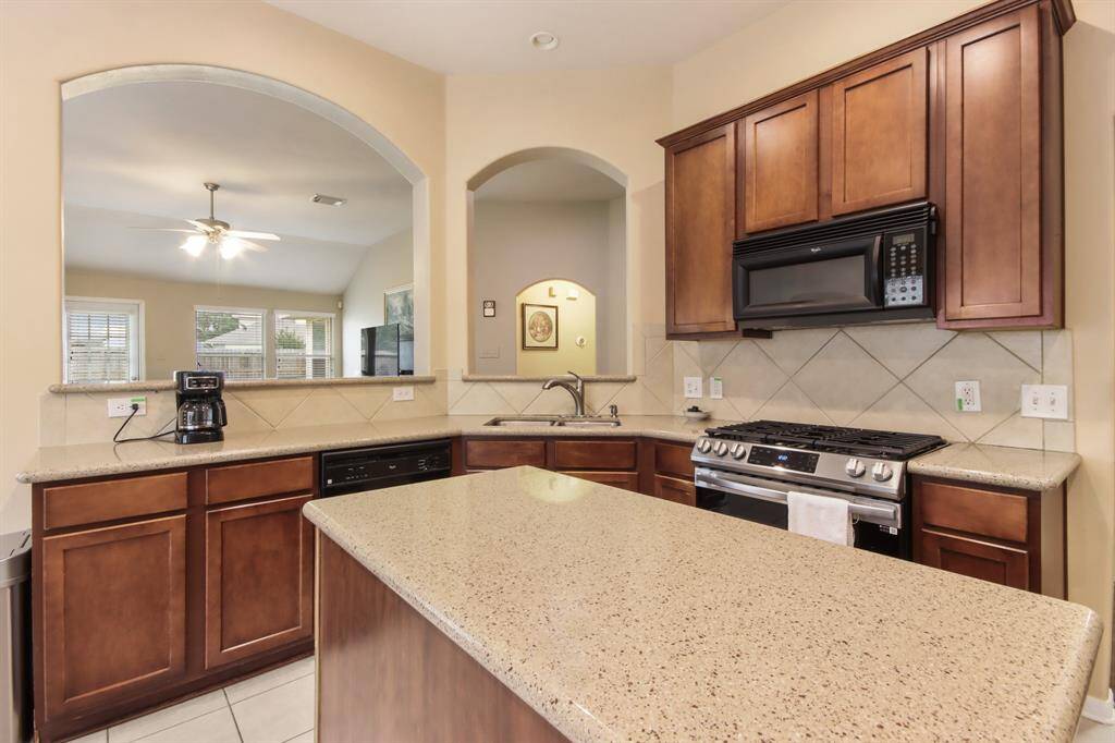 This kitchen features warm wooden cabinets, a spacious island with a speckled countertop, and modern appliances. An open archway connects to a bright living area with large windows and a ceiling fan, creating a welcoming, open-concept space.